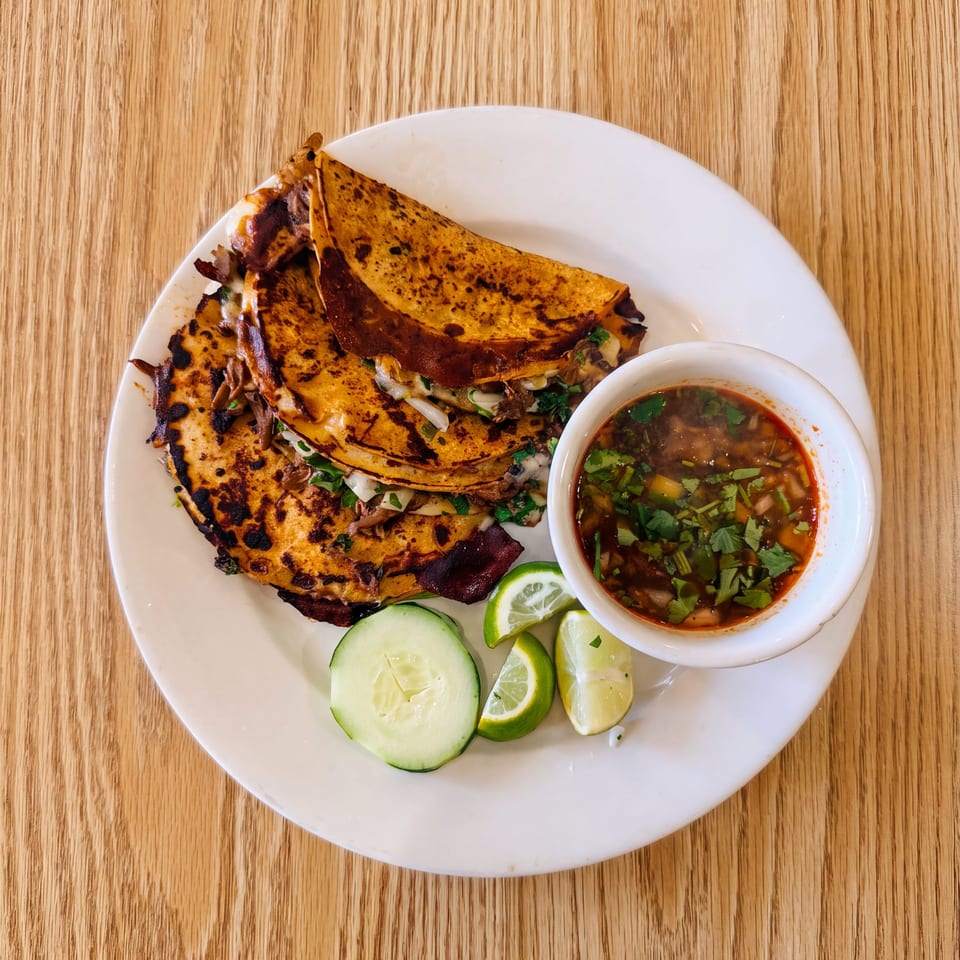 Plate of crispy birria tacos with shredded meat, cilantro, consommé dipping broth, lime wedges, and cucumber slices.