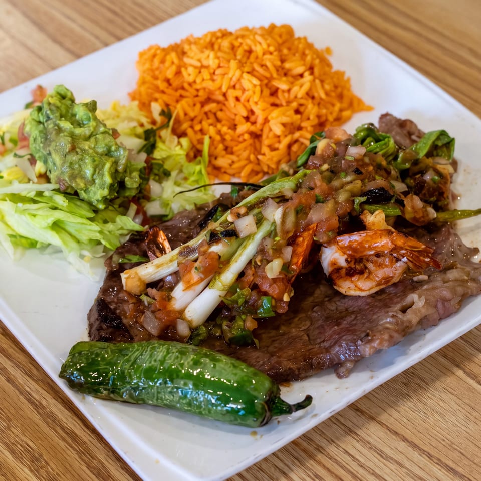 Grilled steak topped with shrimp and vegetables, served with orange rice, guacamole, salad, and a roasted green pepper.