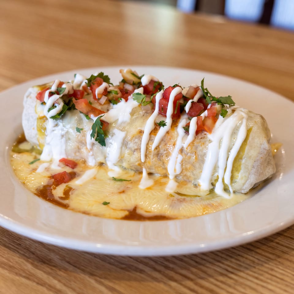 Burrito topped with diced tomatoes, cilantro, and white sauce, served on a plate over melted cheese.