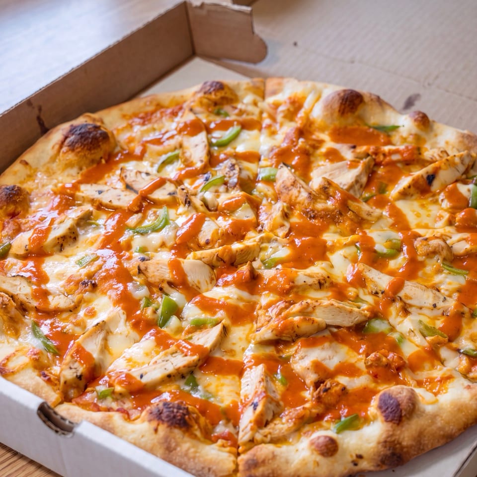 Cheesy chicken pizza topped with green peppers and orange sauce in an open cardboard delivery box.