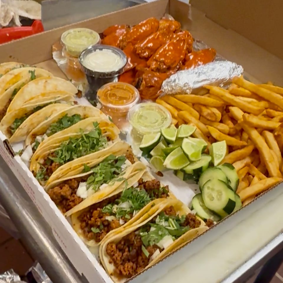 A takeout box filled with tacos, buffalo wings, french fries, and sides of lime, cucumber, and sauces.